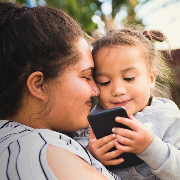 Photo contains a mother nuzzling her young daughter, who is looking at a cell phone amusedly. 