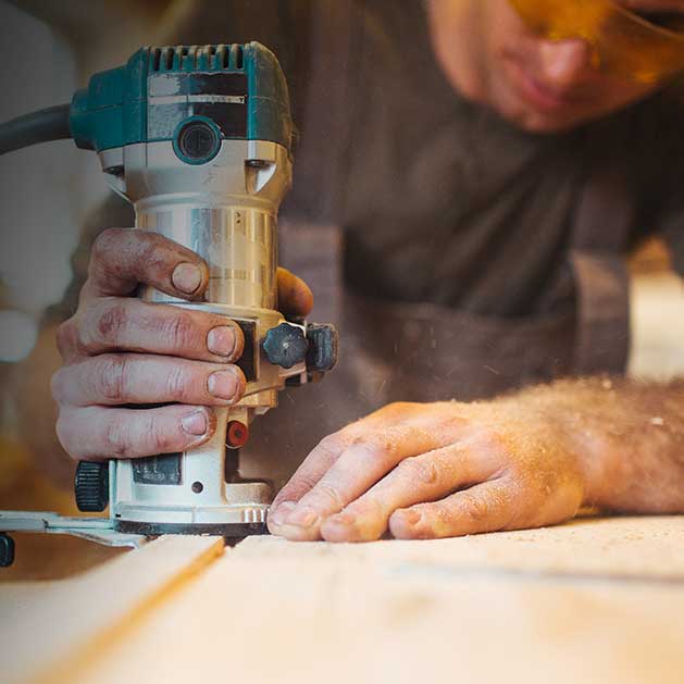 Photo contains a close-up view of a man in safety glasses using a router on a wooden board