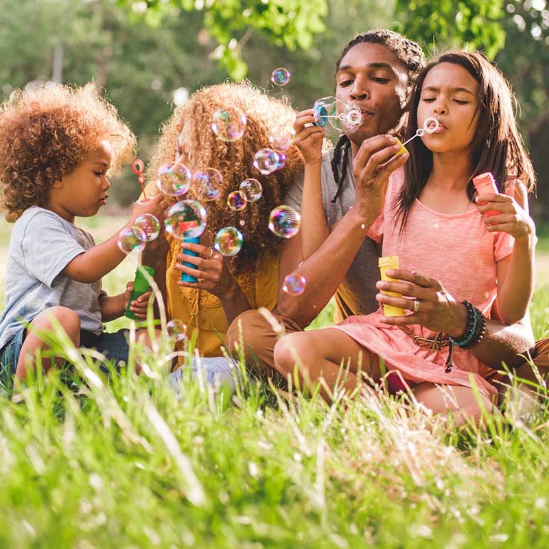 A young, black family, basked in the late afternoon light, blows bubbles in the grassy field of a bucolic community park. 
