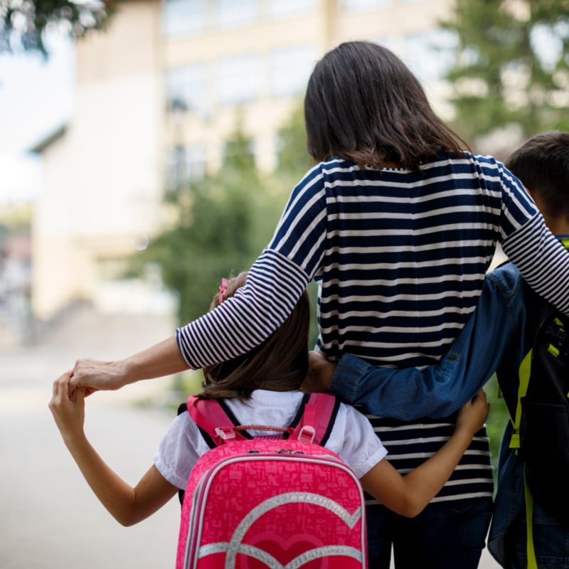Photo of a mother walking a young boy and girl with backpacks to school. Shot from behind